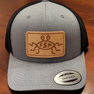 Op de afbeelding: Grijze en zwarte truckerpet met een bruin leren patch met de letters "FSM" en een cartoonafbeelding. De pet heeft een gebogen klep en een snapbacksluiting. Het label luidt "The Authentic Snapback Yupoong Since 1974."