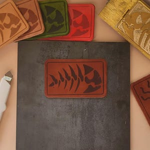 May include: Five leather patches with different designs. The patches are brown, green, red, and a darker brown. The designs include a skull, a fish skeleton, and a stylized fish. The patches are on a dark gray surface.