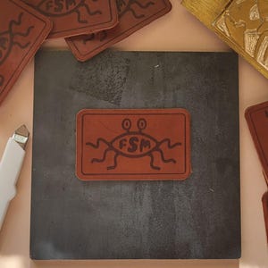 Puede incluir: Un parche de cuero marrón con una criatura alienígena negra con dos ojos y el texto "FSM" dentro de una forma redondeada.
