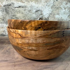 Puede incluir: Dos cuencos de madera apilados. Los cuencos son de madera marrón claro con vetas marrón oscuro y negro. La veta de la madera es visible, creando un aspecto natural y rústico. Están sobre una superficie de madera.