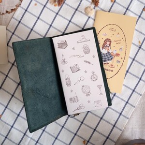May include: A dark green leather journal cover holding a white notebook with black line art illustrations of vintage items like a globe, clock, and candle, alongside a yellow notebook featuring a cartoon illustration and the text "slow living COFFEE days".