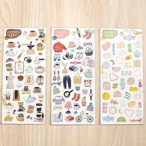 Könnte beinhalten: Drei Stickerbögen mit dem Text "HANKO SEAL". Ein Bogen zeigt Illustrationen zum Thema Kaffee, ein anderer Kleidung und Accessoires, und der dritte abstrakte Formen in Pastellfarben. Die Aufkleber befinden sich auf einem hellen Holzgrund.