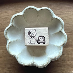 May include: A wooden stamp depicting a cat on the phone, alongside a vintage rotary dial telephone. The stamp is placed on a pale green, scalloped ceramic dish. The dish sits on a wooden surface.