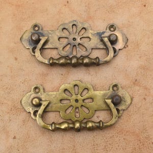 Puede incluir: Dos tiradores de cajón de latón a juego con una placa central en forma de flor de ocho pétalos y un asa curva oscilante. Cada pieza incluye dos orificios de montaje circulares en los extremos para su fijación a muebles o armarios.
