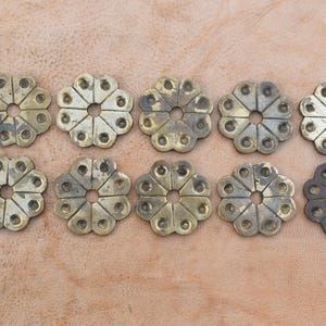 May include: Ten brass-coloured metal decorative pieces with a flower-like design. Each piece has a central hole and twelve points around the edge. One piece is a darker shade of brass.