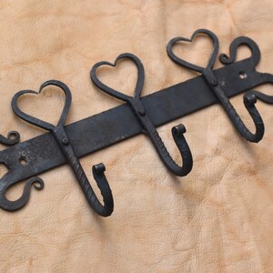 May include: A black metal wall hook with three heart-shaped details. The hooks are curved and the mounting bar has decorative swirls. The piece is set against a light brown background. A decorative item for the home.