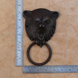 Puede incluir: Un llamador de puerta de cabeza de oso de hierro fundido con un tirador de anillo. El oso tiene una expresión de gruñido y está pintado de color marrón oscuro.