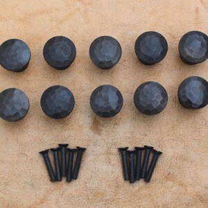 May include: Ten round, dark grey metal cabinet knobs featuring a hammered, textured surface. The set includes ten matching black metal screws with flat heads, arranged in two groups of five below the knobs for installation purposes.