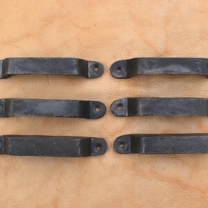 May include: Six black metal handles, each with a rectangular body and rounded ends, featuring two mounting holes. They are arranged in two rows of three on a light brown leather surface. The handles are likely made of cast iron.