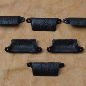 May include: Six black cast iron drawer pulls with a decorative design. The pulls are all the same size and shape, and they have a slightly curved shape.