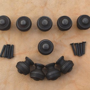 May include: A set of nine matte black cabinet knobs with a rounded, tiered profile. The collection includes five knobs shown from a top-down perspective, four knobs shown from a side profile, and eight matching black metal mounting screws.