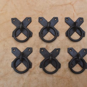 May include: A set of six dark metal drawer pulls, each featuring an X-shaped mounting plate with two screw holes and a circular ring handle attached to the center. The hardware has a matte finish and is arranged in two rows of three identical pieces.