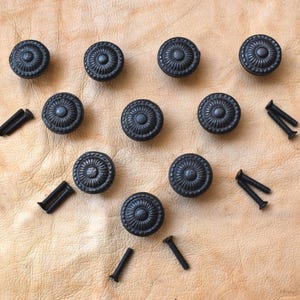 Könnte beinhalten: Zehn schwarze, runde Schrankknöpfe mit detailliertem floralem Design und passenden Schrauben. Sie sind auf einer hellbraunen, strukturierten Oberfläche angeordnet, wahrscheinlich Leder oder einem ähnlichen Material.