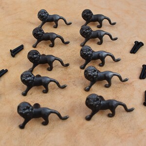 Może przedstawiać: Dziesięć czarnych metalowych gałek do szuflad z głową lwa i śrubami. Głowy lwów są w pozycji chodzenia z zamkniętą paszczą.