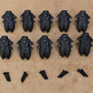 Puede incluir: Diez tiradores de cajón de hierro fundido negro en forma de cigarra con tornillos. Los tiradores están detallados con un diseño de cigarra realista.