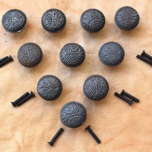 May include: A collection of ten dark gray, round cabinet knobs with an embossed pattern. Each knob is accompanied by a black screw. The knobs are arranged on a light brown surface, likely leather or a similar material.