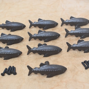 Könnte beinhalten: Zehn schwarze, gusseiserne Fisch-Schubladenknöpfe mit strukturierter Oberfläche. Die Knöpfe sind in drei Reihen angeordnet. Jeder Knopf hat ein Schraubenloch auf der Rückseite. Die Knöpfe werden mit acht schwarzen Schrauben geliefert.