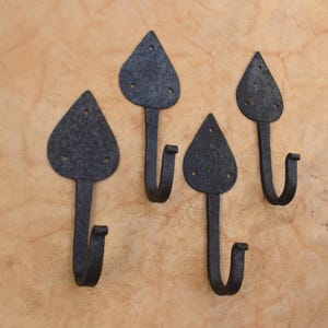 Pode incluir: Um conjunto de quatro ganchos de parede de metal cinza-escuro, cada um com uma placa de montagem texturizada em forma de coração com dois orifícios pré-perfurados no topo e um gancho curvo na parte inferior, dispostos em formação escalonada.