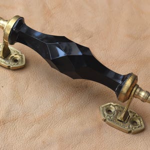 Peut inclure: Une poignée de porte noire à facettes avec des accents et des plaques de montage de couleur laiton. La poignée a un design géométrique et convient aux armoires ou aux portes. La quincaillerie est présentée sur un fond beige.
