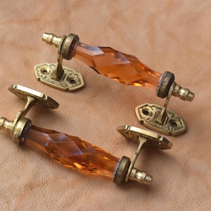 Op de afbeelding: Twee vintage kastgrepen met facetgeslepen amberkleurige glazen handgrepen en sierlijke messing hardware. De grepen zijn geplaatst op een getextureerd, bruin oppervlak, wat hun decoratieve ontwerp en antieke uitstraling benadrukt. Elke greep is ongeveer 15 cm lang.