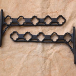 May include: Two black cast iron shelf brackets. Each bracket has a vertical support and a horizontal bar with four clover-shaped cutouts. The brackets are displayed on a textured, tan surface.