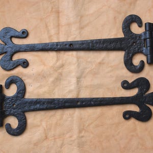 May include: Two black, ornate iron hinges with decorative fleur-de-lis and scrollwork designs. The hinges are laid out on a light brown, textured surface, showcasing their detailed craftsmanship and vintage aesthetic.