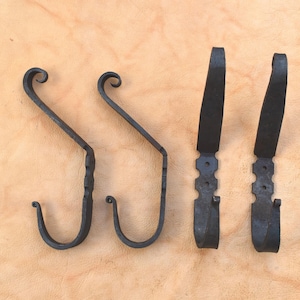 Pode incluir: Quatro ganchos de ferro forjado preto. Dois têm formato de S com a parte superior enrolada e dois são planos com uma base decorativa. Os ganchos são exibidos em uma superfície marrom clara.