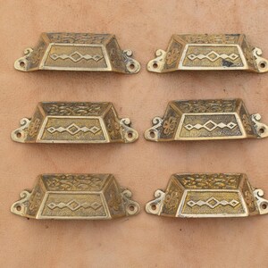 May include: A set of six brass drawer pulls arranged in two columns of three. Each pull features a trapezoidal shape with an embossed geometric diamond pattern on the front face and decorative scrollwork details on the mounting flanges.