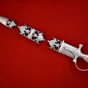 Può includere: Un pugnale d'argento con intricati disegni floreali, diviso in tre pezzi. Il pugnale è su una superficie rossa.