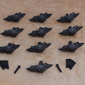 May include: A collection of ten black metal cabinet pulls with a winged design, along with a set of screws. The pulls are arranged in a grid pattern on a textured, tan surface. The hardware is likely for furniture.