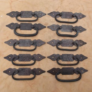 May include: A set of ten dark gray metal drawer pulls arranged in two columns. Each pull features a rectangular backplate with pointed ends and a rounded, swinging handle attached to two circular mounting points on the plate surface.