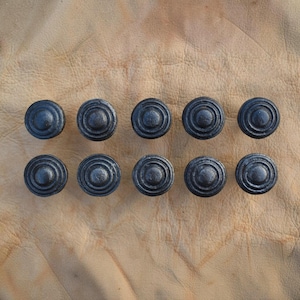 May include: Ten circular black metal knobs arranged in two rows of five. Each knob features a raised center point surrounded by three concentric circular ridges, creating a textured, stepped profile on the face of the hardware pieces.