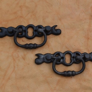 Puede incluir: Dos tiradores de cajón de metal negro con un diseño decorativo. Cada tirador tiene una manija de anillo y una placa posterior decorativa.