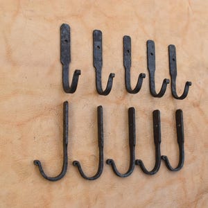 Pode incluir: Um conjunto de dez ganchos de parede de metal cinza-escuro dispostos em duas filas de cinco. Cada gancho possui uma placa de montagem vertical plana com um orifício circular para parafuso no topo e um gancho curvo em forma de J na base.