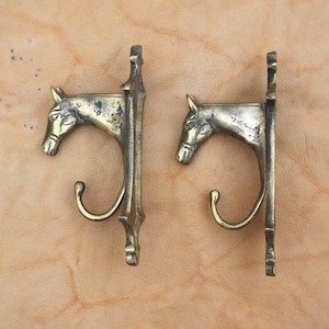 May include: A pair of antique brass wall hooks. Each hook is shaped like a horse's head with a curved hook beneath. The hooks are mounted on a rectangular backplate. The brass has an aged, tarnished appearance.