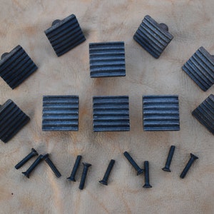 May include: Ten black metal cabinet knobs with a ribbed design and a square shape. The knobs are arranged in a semi-circle with screws scattered around them.