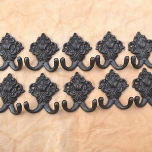 Pode incluir: Dez cabides duplos pretos e ornamentados com um design de flor-de-lis. Os ganchos são feitos de ferro fundido e estão dispostos em duas filas de cinco contra um fundo castanho claro. Cada gancho tem um topo decorativo.