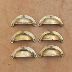 Può includere: Un set di sei maniglie a conchiglia in ottone a forma di semicerchio, disposte in due colonne da tre. Ogni maniglia presenta un design curvo e liscio con due piccoli fori circolari per le viti alle estremità per il fissaggio.