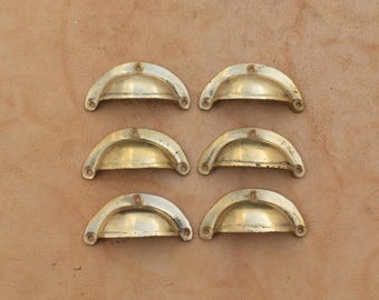 May include: A set of six semi-circular brass cup drawer pulls arranged in two columns of three. Each pull features a smooth, curved handle design with two small circular screw holes located at the top and bottom ends for mounting purposes.