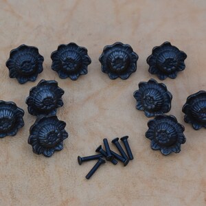 May include: Ten black cast iron drawer pulls with a floral design. The pulls are arranged in a circle with a pile of black screws in the centre.