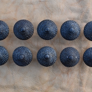 May include: Ten black metal cabinet knobs with an ornate design. The knobs have a raised centre and a decorative pattern around the edge.