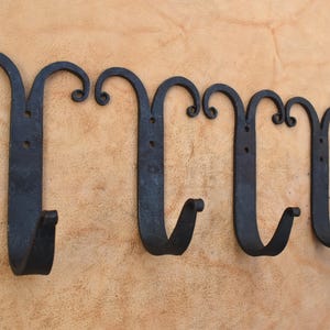 Pode incluir: Um conjunto de quatro ganchos de parede em metal preto, cada um com uma barra plana vertical, um gancho em forma de J na parte inferior e dois ornamentos simétricos em forma de voluta no topo, com dois orifícios de montagem circulares.