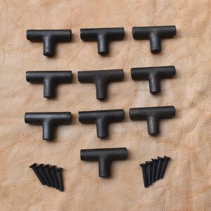 May include: A collection of ten black, T-shaped cabinet pulls with matching screws. The hardware is made of a dark metal and is arranged on a tan leather surface. The pulls are likely for drawers or cabinets.