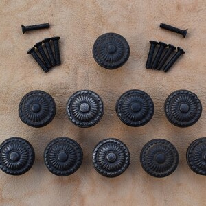 May include: Ten black metal cabinet knobs with a decorative floral design. The knobs are arranged in a pyramid shape with screws scattered around them.