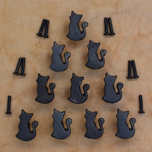 Könnte beinhalten: Ein Satz von zehn schwarzen, katzenförmigen Schubladenknöpfen aus Metall mit Schrauben. Die Knöpfe sind in Dreiecksform auf einer hellbraunen Oberfläche angeordnet.