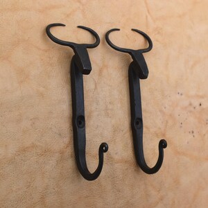 Pode incluir: Dois ganchos de parede em metal preto com um design estilizado de cabeça de touro no topo. Cada gancho tem uma secção inferior curva para pendurar objetos e um orifício de montagem central. Acabamento escuro, mate e texturizado.