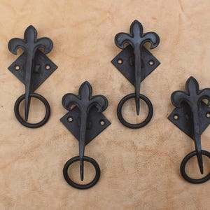 May include: A set of four black metal drawer pulls, each featuring a diamond-shaped backplate with two screw holes, a fleur-de-lis top ornament, and a circular drop ring handle attached to the center, arranged in a staggered formation.