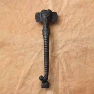 May include: A black, decorative door handle shaped like an elephant. The handle features an elephant head with detailed carvings, a long trunk, and a textured body. The handle is made of a dark material and is set against a light brown background.