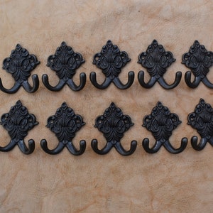 May include: Ten black metal double hooks with ornate scrollwork designs. The hooks are mounted on a light brown surface.