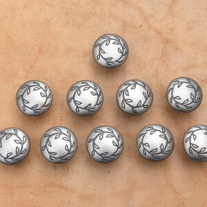 May include: A collection of ten circular silver-toned metal buttons arranged in a triangular formation. Each button features a raised, engraved circular border design consisting of delicate, interconnected leafy vine patterns surrounding a smooth centre.